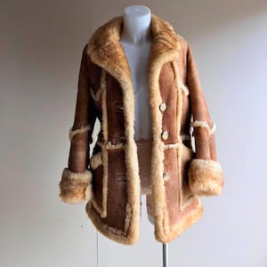 May include: A brown vintage coat with a tan shearling lining and trim. The coat features a notched collar, button closures, and fur cuffs. The coat is displayed on a mannequin.