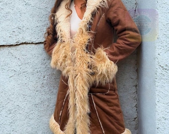 May include: A brown suede coat with cream-colored faux fur trim. The coat features a longline silhouette, a notched collar, and fur cuffs. The coat has a vintage aesthetic and is buttoned up. The coat is a warm outerwear garment.
