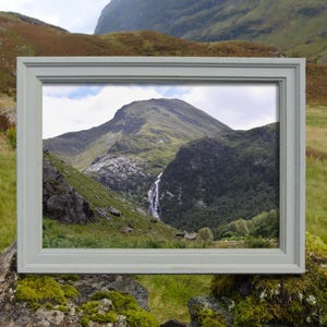 May include: A framed landscape photograph of a mountain scene with a waterfall. The frame is a light gray color, and the photograph shows a lush green mountain with a waterfall cascading down its side. The scene is set in a natural environment.