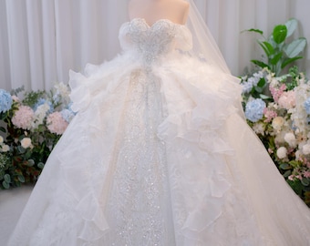 Può includere: Un abito da ballo bianco senza spalline con scollo a cuore, intricati decori di paillettes argentate e voluminosi strati di volant. L'abito include un lungo velo trasparente e una piccola corona dorata posizionata sul manichino.