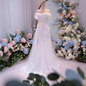 May include: A white wedding dress on a mannequin, featuring an off-the-shoulder neckline and floral embellishments. The dress is displayed in front of a large mirror and surrounded by floral arrangements in pastel colors, creating an elegant bridal theme.