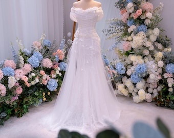 Può includere: Abito da sposa bianco su manichino, con scollatura a spalle scoperte e decorazioni floreali. L'abito è esposto davanti a un grande specchio e circondato da composizioni floreali dai colori pastello, creando un tema nuziale elegante.