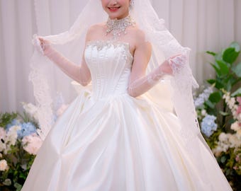 Può includere: Un abito da ballo bianco senza spalline con un corpetto in stile corsetto caratterizzato da decorazioni argentate sullo scollo. L'abito include un'ampia gonna plissettata, guanti in pizzo trasparente e un lungo velo coordinato bordato di pizzo.