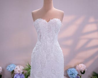 Può includere: Un abito corto bianco senza spalline caratterizzato da intricati ricami in pizzo e decorazioni con paillettes scintillanti su tutto il tessuto. Il capo è esposto su un manichino sormontato da un accessorio a corona in filo dorato.
