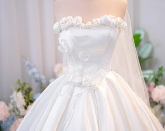 Può includere: Un abito da sposa bianco senza spalline con scollo a cuore decorato con rose in tessuto, un corpetto aderente con ulteriori accenti floreali in vita, una gonna ampia e un velo trasparente sormontato da una corona in metallo dorato.