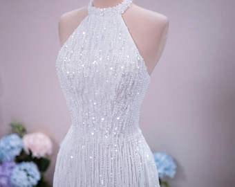 Può includere: Un abito bianco senza maniche con scollo all'americana, caratterizzato da fitte file verticali di perline e paillettes scintillanti che creano un effetto frangiato. L'indumento è esposto su un manichino che indossa una corona in metallo dorato.