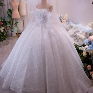 Op de afbeelding: Een witte trouwjurk met een sprankelend lijfje en een volle, gelaagde rok. Het off-the-shoulder ontwerp is versierd met delicate kant en tule. Een gouden kroon en sluier worden op een mannequin getoond. Het woord "Chi" is zichtbaar.