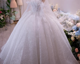 Può includere: Un abito da ballo bianco senza spalline con corpetto a corsetto, caratterizzato da intricati decori di paillettes argentate su tutta l'ampia gonna e un velo trasparente abbinato con una corona in metallo dorato su un manichino.