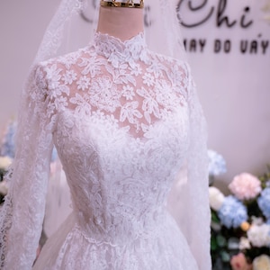 May include: A white long-sleeved lace gown featuring a high neckline with intricate floral embroidery and a sheer lace bodice. The dress is displayed on a mannequin wearing a gold crown-shaped headpiece and a long, sheer white lace veil.