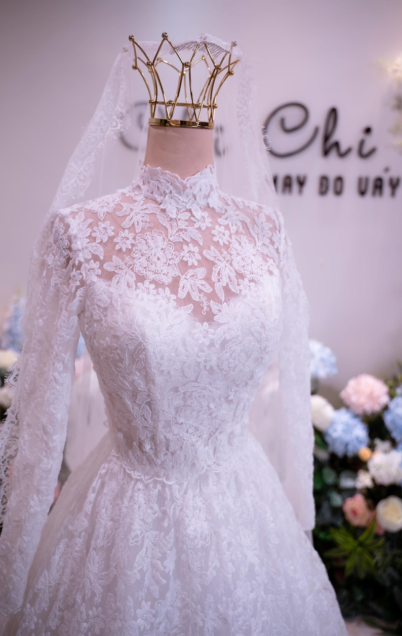 May include: A white long-sleeved lace gown featuring a high neckline with intricate floral embroidery and a sheer lace bodice. The dress is displayed on a mannequin wearing a gold crown-shaped headpiece and a long, sheer white lace veil.