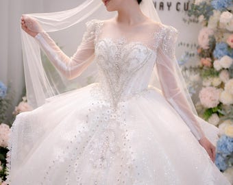 Può includere: Un abito da sposa bianco a palloncino con maniche lunghe trasparenti, scollo a cuore e un corpetto intricato decorato con ricami argentati e paillettes. L'ampia gonna è composta da strati di tulle, decorazioni scintillanti e pizzo.