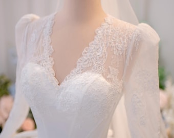 Può includere: Un abito bianco a maniche lunghe con scollo a V e dettagli in pizzo intricati sul corpetto e sulle maniche, abbinato a un velo trasparente e a una corona in metallo dorato posizionata sulla testa del manichino.