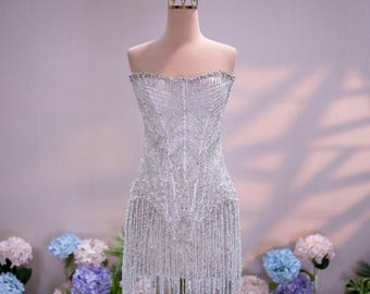 Può includere: Un abito corto senza spalline ricoperto di paillettes argentate e intricate perline, caratterizzato da un corpetto aderente e un orlo rifinito con lunghe frange di perline pendenti che creano un effetto scintillante sull'intera superficie.