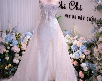 May include: A white wedding dress on a mannequin, featuring a beaded bodice and a flowing skirt with a high slit. The dress is adorned with sparkling embellishments. A gold crown and a choker necklace complete the bridal look. The background includes floral arrangements and the text "Thi Chi".