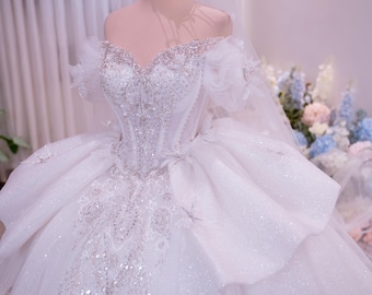 Può includere: Un abito da ballo bianco con spalle scoperte, scollo a cuore, intricate perline argentate e decorazioni di paillettes. L'abito presenta gonne voluminose a strati con una finitura scintillante, abbinato a un velo trasparente e una corona dorata.