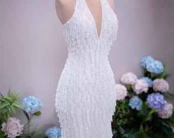 Può includere: Un abito bianco lungo fino ai piedi con scollo all'americana, profondo scollo a V e una texture di frange di perline su tutta la superficie che crea un effetto scintillante. L'abito è esposto su un manichino con una corona in filo dorato.