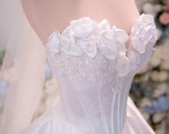 Può includere: Un abito da sposa bianco senza spalline con un corpetto in stile corsetto con stecche verticali, decorato con fiori in tessuto tridimensionali e piccole perle bianche lungo lo scollo, che prosegue in una gonna in raso liscia e ampia.