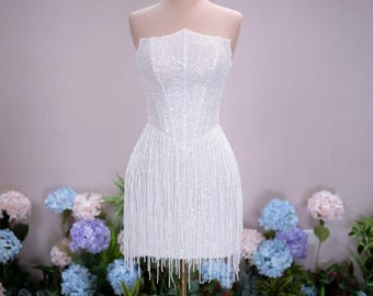 Può includere: Un abito corto bianco senza spalline con corpetto ricoperto di fitte paillettes e perline bianche, e una gonna composta da lunghe frange di perline bianche pendenti. Un copricapo dorato a forma di corona poggia sul collo del manichino.