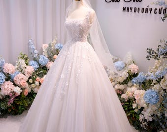 Può includere: Un abito da sposa bianco lungo fino a terra con scollo quadrato e spalline larghe, caratterizzato da un intricato ricamo floreale in pizzo sul corpetto che scende sulla gonna in tulle, abbinato a un velo leggero e trasparente.