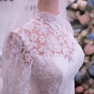 May include: A white long-sleeved wedding dress featuring an intricate floral lace overlay, a high neckline, and a sheer lace bodice. The garment is displayed on a mannequin with a matching lace veil draped over the head, showcasing detailed embroidery.