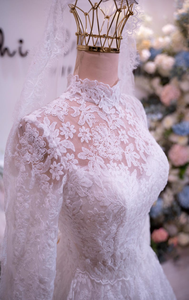 May include: A white long-sleeved wedding dress featuring an intricate floral lace overlay, a high neckline, and a sheer lace bodice. The garment is displayed on a mannequin with a matching lace veil draped over the head, showcasing detailed embroidery.