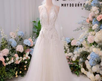 Può includere: Un abito da sposa bianco lungo fino ai piedi con profondo scollo a V, intricati ricami argentati e decorazioni di paillettes. Il corpetto presenta un design all'americana con inserti trasparenti e una gonna in tulle a più strati.