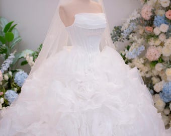 Può includere: Un abito da sposa bianco senza spalline con un corpetto strutturato e una gonna voluminosa composta da strati di tessuto arricciato. Un velo trasparente che arriva a terra è drappeggiato sopra e una piccola corona in filo dorato è sul manichino.