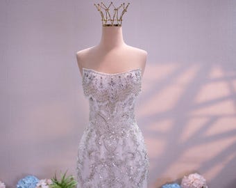 Può includere: Un abito corto senza spalline di colore bianco con intricate perline argentate e ricami di paillettes in un motivo a volute. Il capo è esposto su un manichino sormontato da una corona in filo dorato, mostrando una texture densa e scintillante.