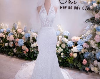 Può includere: Abito da sposa bianco, lungo fino al pavimento, con silhouette a sirena, esposto su un manichino. L'abito presenta uno scollo all'americana e dettagli in pizzo intricati. Una corona e un velo sono sulla testa del manichino. Lo sfondo include composizioni floreali e il testo "Chi MAY DO VAY CUOI".