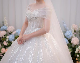 Può includere: Un abito da sposa bianco a palloncino con spalle scoperte, scollo a cuore, corpetto aderente con intricati decori di paillettes argentate e una gonna voluminosa lunga fino a terra ricoperta di paillettes scintillanti con un velo trasparente.