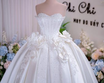 Può includere: Un abito da ballo bianco senza spalline con scollo a cuore decorato con perline, un corpetto in stile corsetto e grandi rose in tessuto in vita. L'ampia gonna presenta delicati ricami floreali e cristalli sparsi su tutta la superficie.