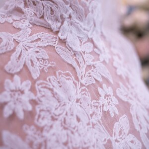 May include: A high-neck garment featuring intricate white floral embroidery layered over a sheer, light-colored mesh fabric. The design showcases detailed stitching patterns of leaves and blossoms that extend from the collar down the bodice.
