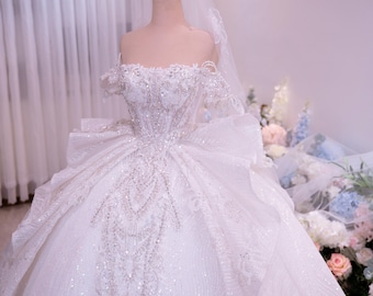Può includere: Un abito da ballo bianco con spalle scoperte caratterizzato da intricate perline e paillettes argentate sul corpetto e sulla gonna. L'abito presenta una gonna voluminosa a strati con strascico, abbinata a un velo trasparente e una corona dorata.