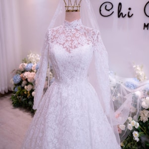 May include: A white floor-length wedding dress featuring long sleeves, a high neckline, and intricate floral lace detailing throughout the bodice and skirt. The gown is paired with a sheer, lightweight veil that drapes over the shoulders.