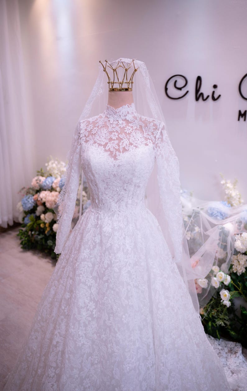 May include: A white floor-length wedding dress featuring long sleeves, a high neckline, and intricate floral lace detailing throughout the bodice and skirt. The gown is paired with a sheer, lightweight veil that drapes over the shoulders.