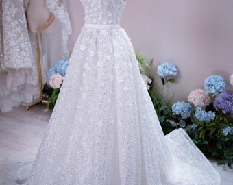 Può includere: Un abito da sposa bianco senza spalline con scollo a cuore, corpetto aderente decorato con piccole applicazioni floreali, vita definita da una fascia sottile e gonna lunga a trapezio ricoperta di paillettes scintillanti e dettagli floreali.