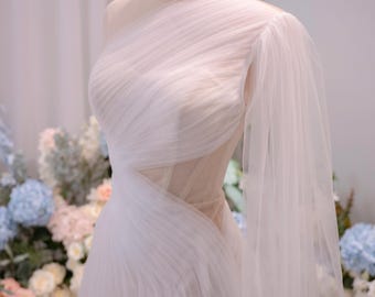May include: A white one-shoulder gown featuring layered tulle fabric with diagonal pleating across the bodice and a sheer side cutout. The dress includes a long, flowing sleeve and is displayed on a mannequin wearing a gold wire crown.
