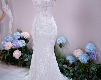 Può includere: Un abito lungo in pizzo bianco con scollo alto, corpetto aderente con intricate perline e silhouette a sirena. L'abito è esposto su un manichino che indossa una piccola corona dorata, mettendo in risalto il dettagliato ricamo floreale.