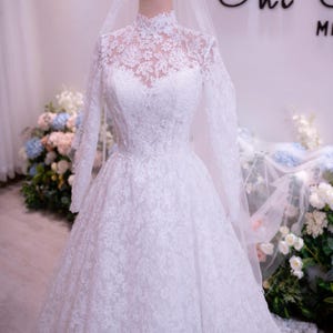 May include: A white floor-length gown featuring long sleeves, a high neckline, and intricate floral lace detailing throughout the bodice and skirt. A sheer veil is draped over the top, and a gold-colored crown sits atop the mannequin head.