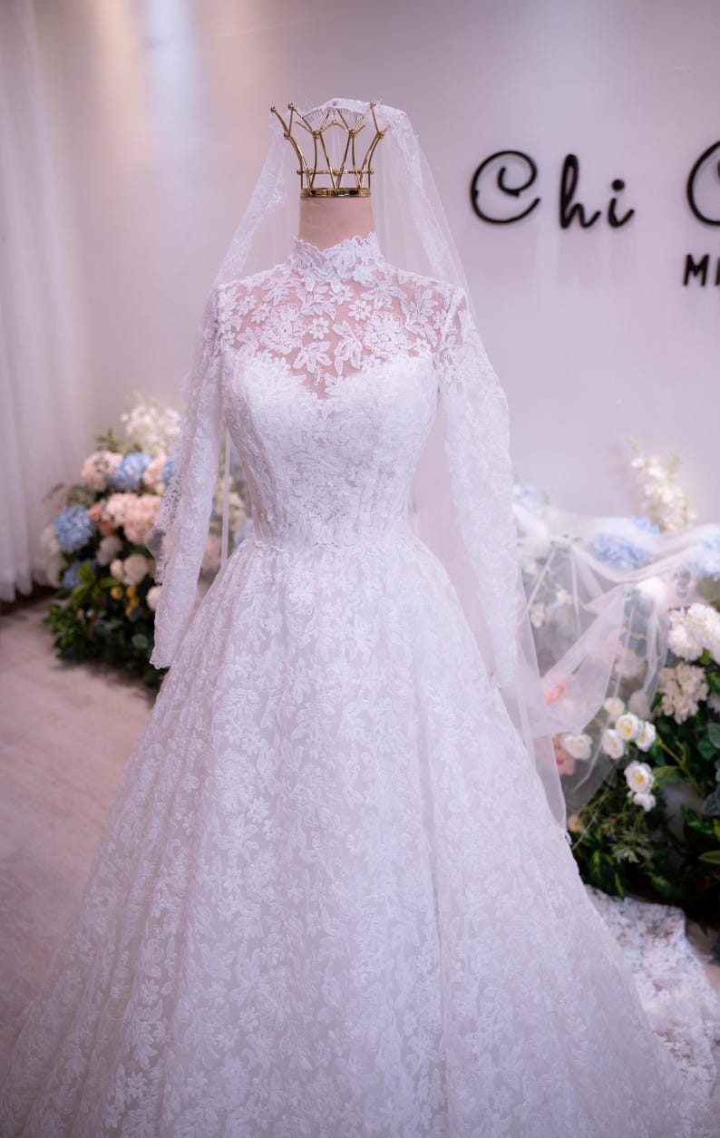 May include: A white floor-length gown featuring long sleeves, a high neckline, and intricate floral lace detailing throughout the bodice and skirt. A sheer veil is draped over the top, and a gold-colored crown sits atop the mannequin head.