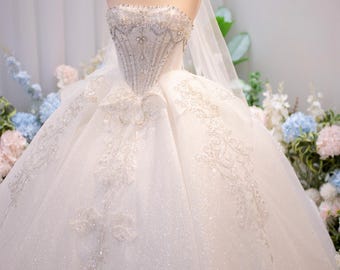 Può includere: Un abito da ballo bianco senza spalline con un corpetto in stile corsetto decorato con intricate perline e ricami argentati. L'ampia gonna è realizzata in tulle glitterato con applicazioni in pizzo floreale e un lungo velo trasparente.