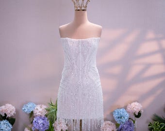 Può includere: Un abito corto bianco senza spalline caratterizzato da un intricato motivo di paillettes e perline argentate, con un orlo a frange. L'abito è esposto su un manichino che indossa una corona di filo dorato, mostrando una silhouette aderente.