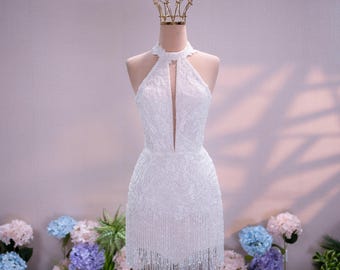 Può includere: Un abito corto bianco senza maniche con scollo all'americana alto e apertura centrale, decorato con intricate perline su tutto il corpetto e un orlo rifinito con lunghe frange pendenti. Il capo è esposto su un manichino con una corona.