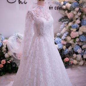 May include: A white floor-length wedding dress featuring long sleeves, a high lace neckline, and an intricate floral lace overlay. The gown is displayed on a mannequin wearing a gold crown-shaped headpiece and a sheer white veil.