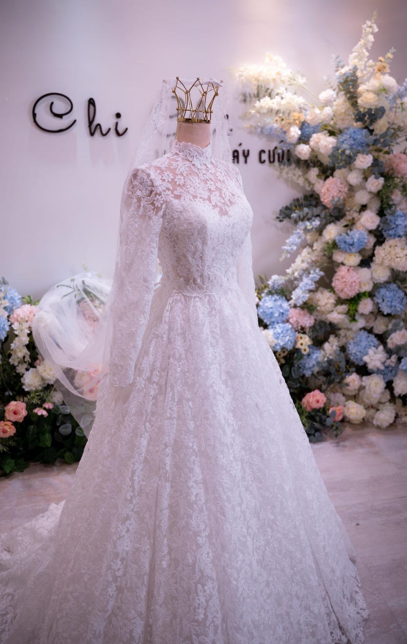May include: A white floor-length wedding dress featuring long sleeves, a high lace neckline, and an intricate floral lace overlay. The gown is displayed on a mannequin wearing a gold crown-shaped headpiece and a sheer white veil.