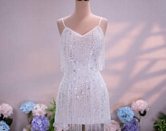 Può includere: Un mini abito bianco con scollo a V, spalline sottili e un design all-over di paillettes scintillanti e lunghe frange pendenti. Il capo è esposto su un manichino sormontato da una piccola corona in filo metallico color oro.