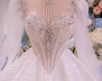 Può includere: Un abito bianco lungo fino ai piedi con scollo alto in pizzo, maniche lunghe trasparenti e un corpetto in stile corsetto decorato con intricate perline argentate, cristalli e perle. L'ampia gonna è realizzata in tessuto scintillante e testurizzato.