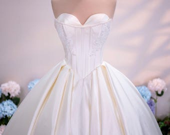Può includere: Un abito da ballo color avorio senza spalline con un corpetto a corsetto strutturato con scollo a cuore, delicati ricami in pizzo bianco e un'ampia gonna in raso plissettato. Una sottile corona in filo dorato poggia sul collo del manichino.