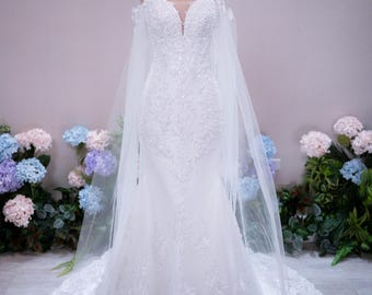 Può includere: Un abito da sposa bianco lungo fino a terra con scollo a cuore, spalline sottili e un intricato ricamo in pizzo su tutto il corpetto e la gonna. L'abito presenta maniche trasparenti scoperte sulle spalle e un lungo mantello in tulle.