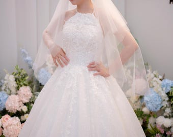 Può includere: Un abito da sposa bianco lungo fino ai piedi con scollo all'americana, intricati ricami in pizzo e un'ampia gonna in tulle. L'abito presenta sottili decorazioni con paillettes ed è abbinato a un velo in tulle trasparente a più strati.